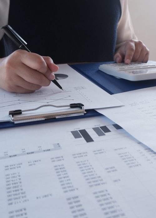 Female working in office, studying using calculator and writing something with documents and chart on table.